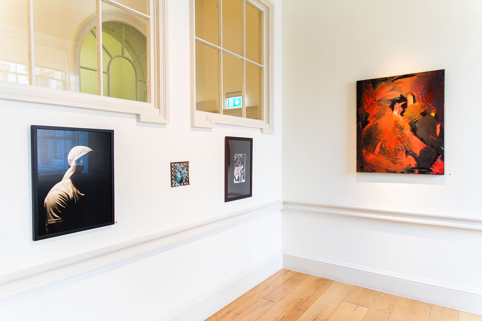 View of the corner of a gallery room. Two white walls, on the right side 3 artworks are hanging, a photograph of a dark skinned person with white clothes against a white background, a blue and brown ceramic piece depicting a face and a black and white photograph. On the right wall, an abstract painting in shades of red.