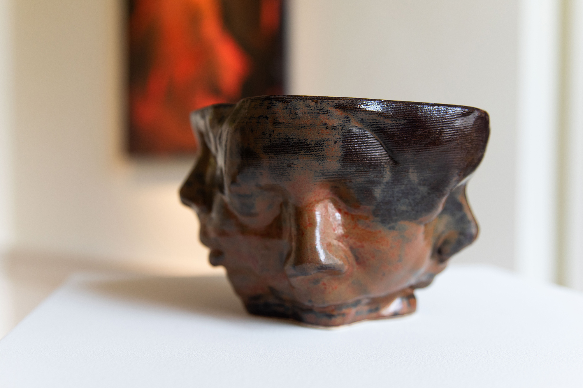 Close-up of brown and red ceramic vessel, on a white plinth. It has faces with closed eyes on it.