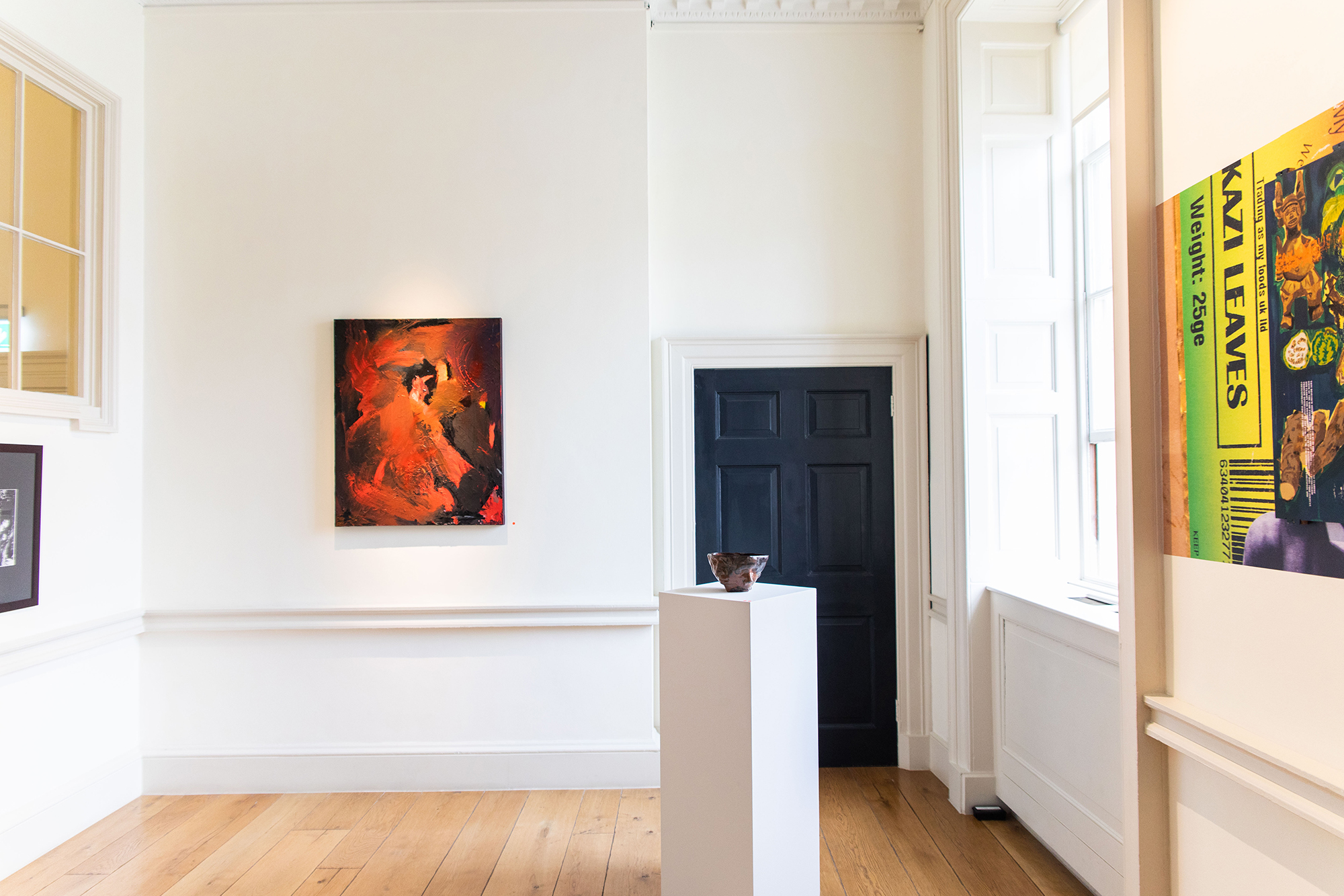 Gallery room showing a painting with abstract red shapes at the far back wall, a ceramic vessel on top of a white plinth and in front of a black door, and a half-cut out of the frame collage on the right.