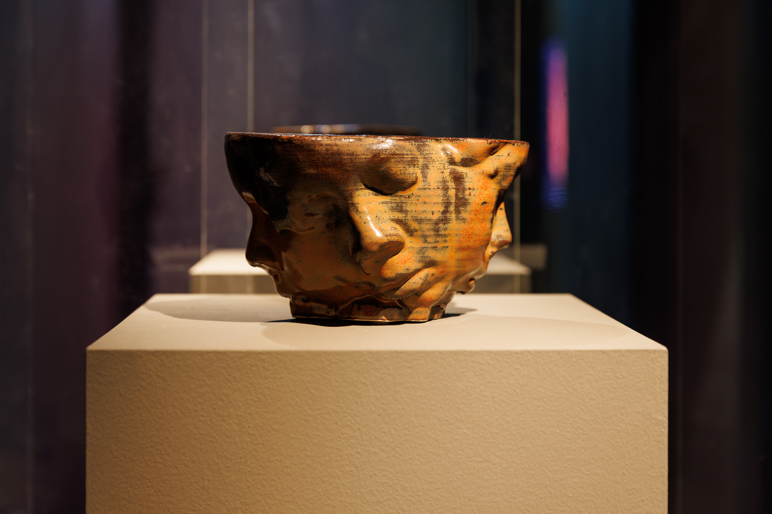 Brown and red glazed ceramic piece on top of a plinth, in a dark room, depicting faces with their eyes closed.
