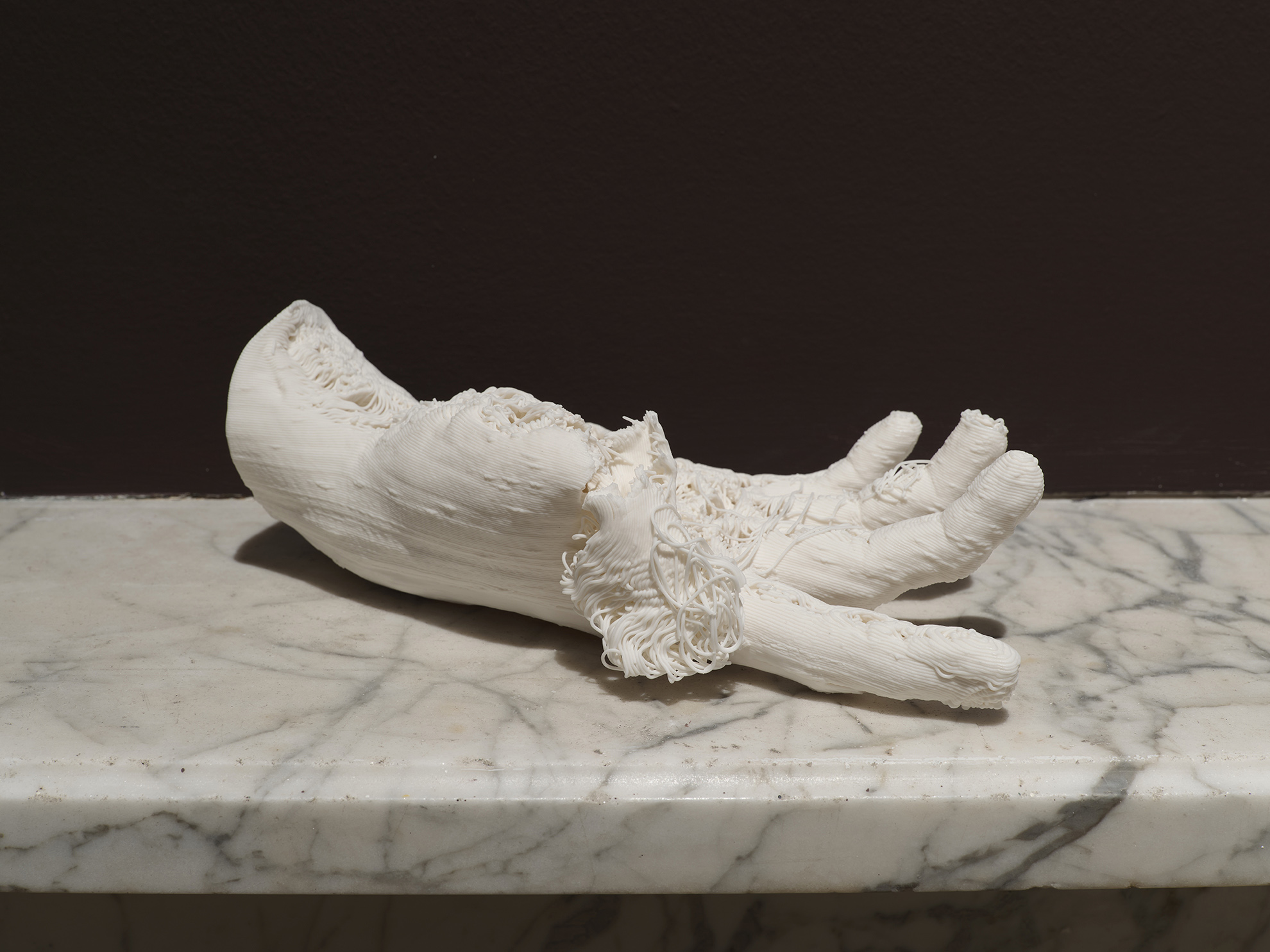 White porcelain hand, 3D printed with errors and torn at the side, on top of a marble mantlepiece against a brown wall.