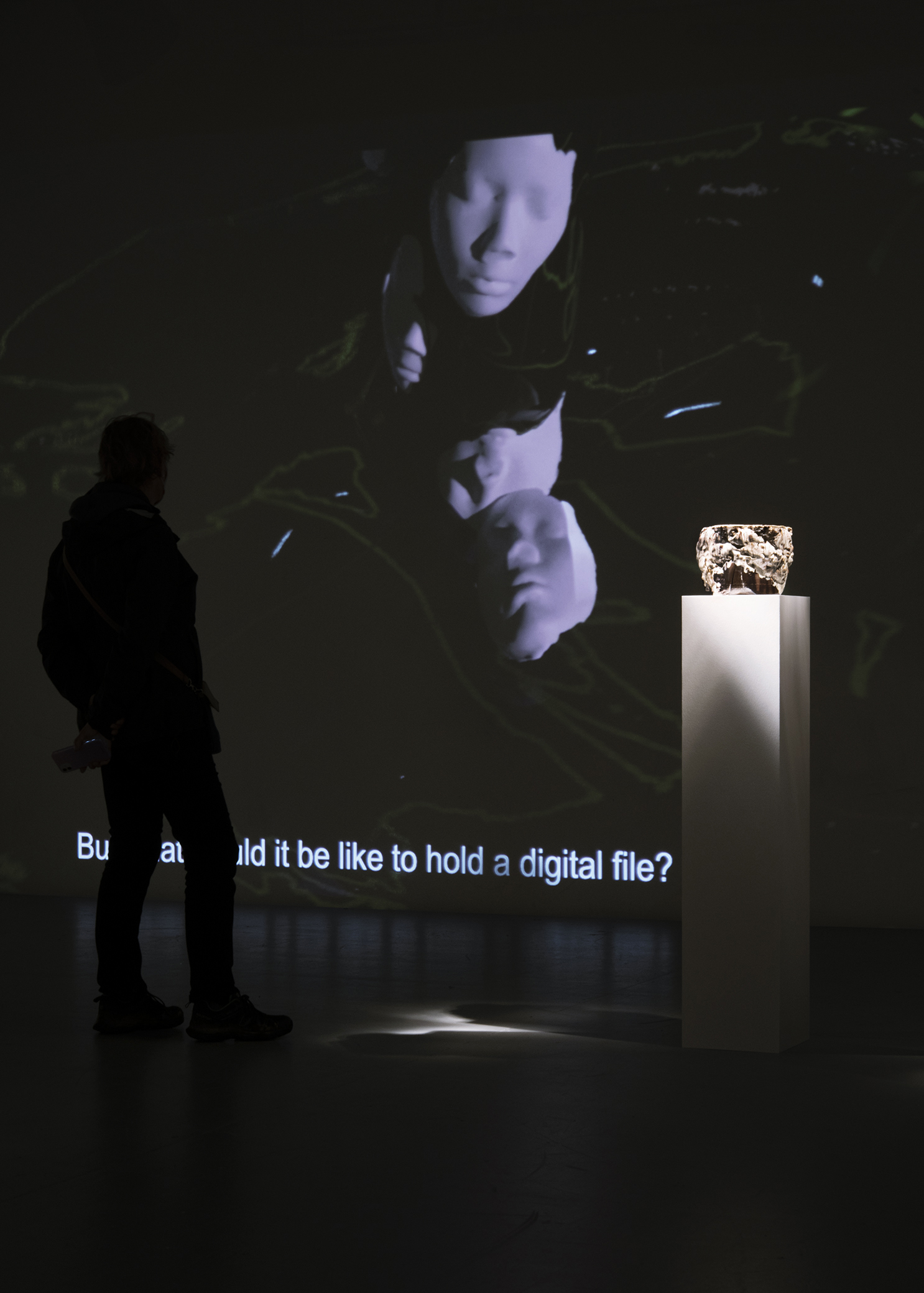 Brown ceramic vessel on a white plinth in a dark room, next to a projection on the wall on the left showing white plaster faces and the text But what would it be like to hold a digital file, but the first part of the text is hidden by the shadow of a person.