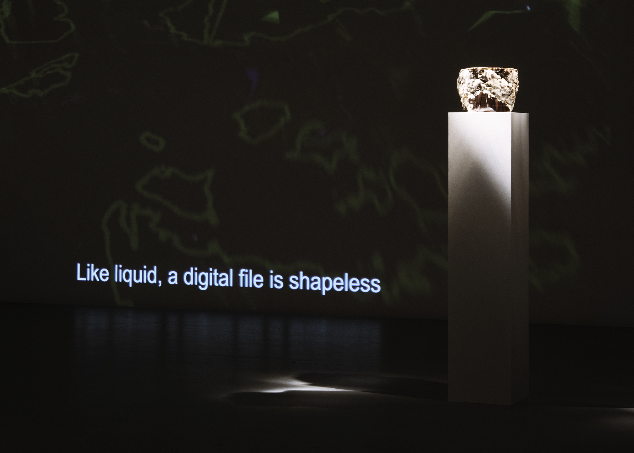 Brown ceramic vessel on a plint in a dark room, next to a projection that shows the words Like liquid, a digital file is shapeless.