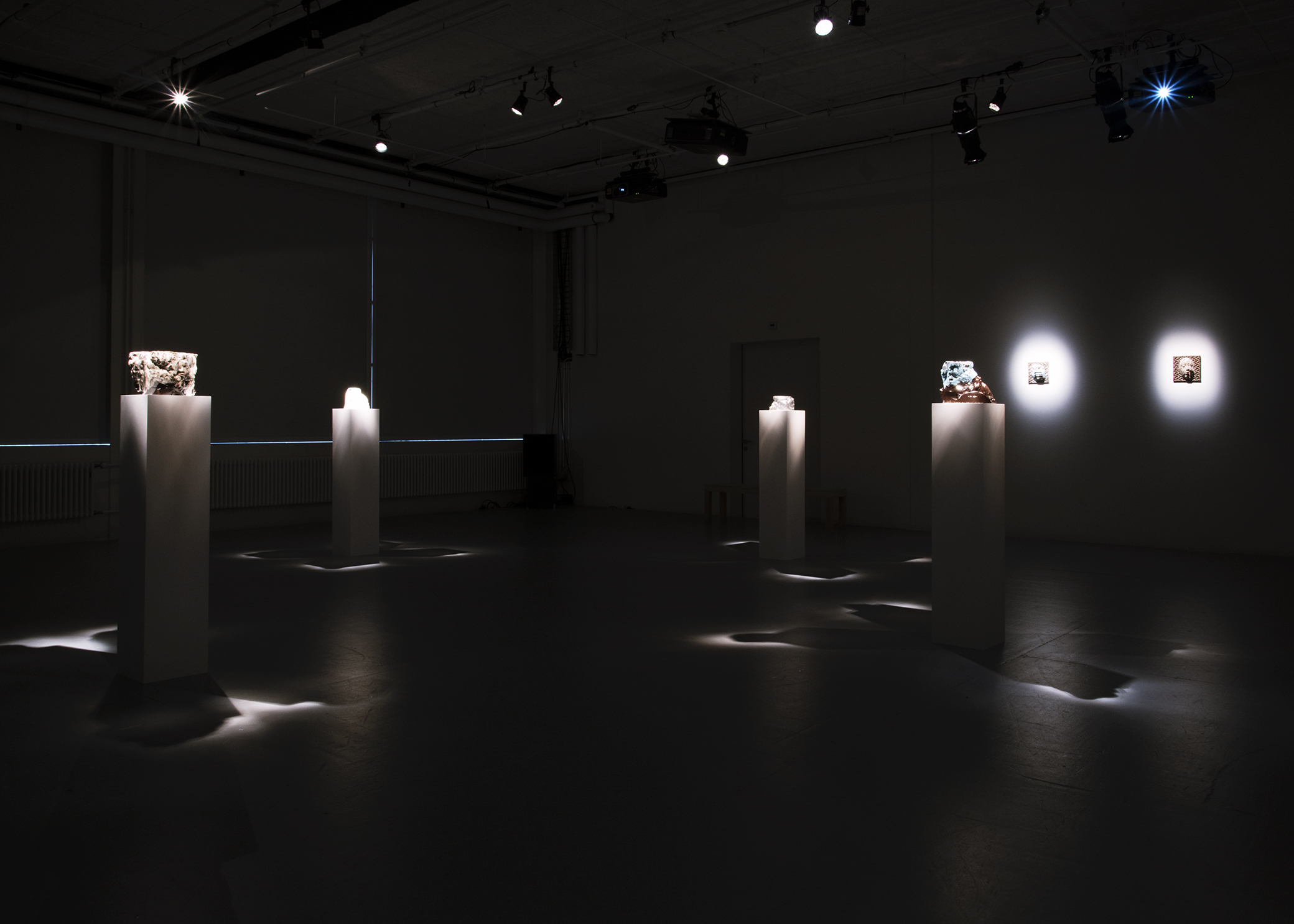 Dark gallery room with multiple ceramic vessels on plinths and two ceramic pieces hung on the wall, all with spotlights on them.