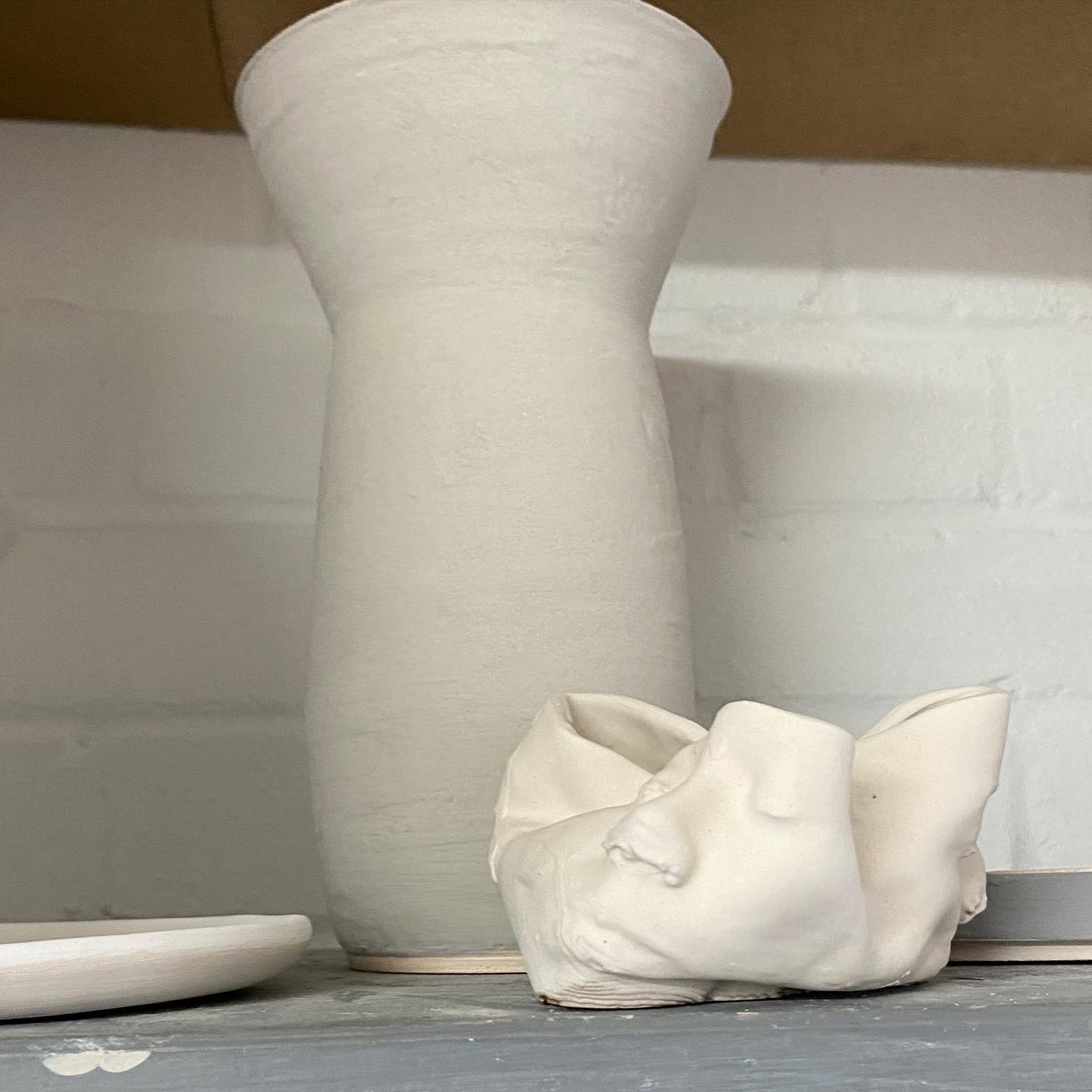 photo of a shelf with three unfired ceramic pieces, all white, a plate on the left side, a tall vase in the back a small ceramic printed vase in the front, as if it was folded, showing a face with closed eyes