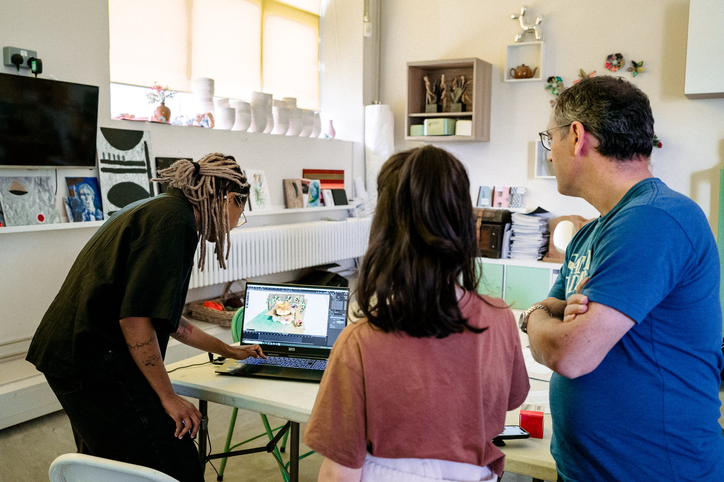 photo displaying three people looking at a laptop in a workshop space. On the left side is the artist, a black person with dreadlocs and black clothing with their hand on the keyboard showing a 3D render to the other two people, a young person with long hair and pink shirt, and their parent, in a blue shirt and short hair with their arms crossed.