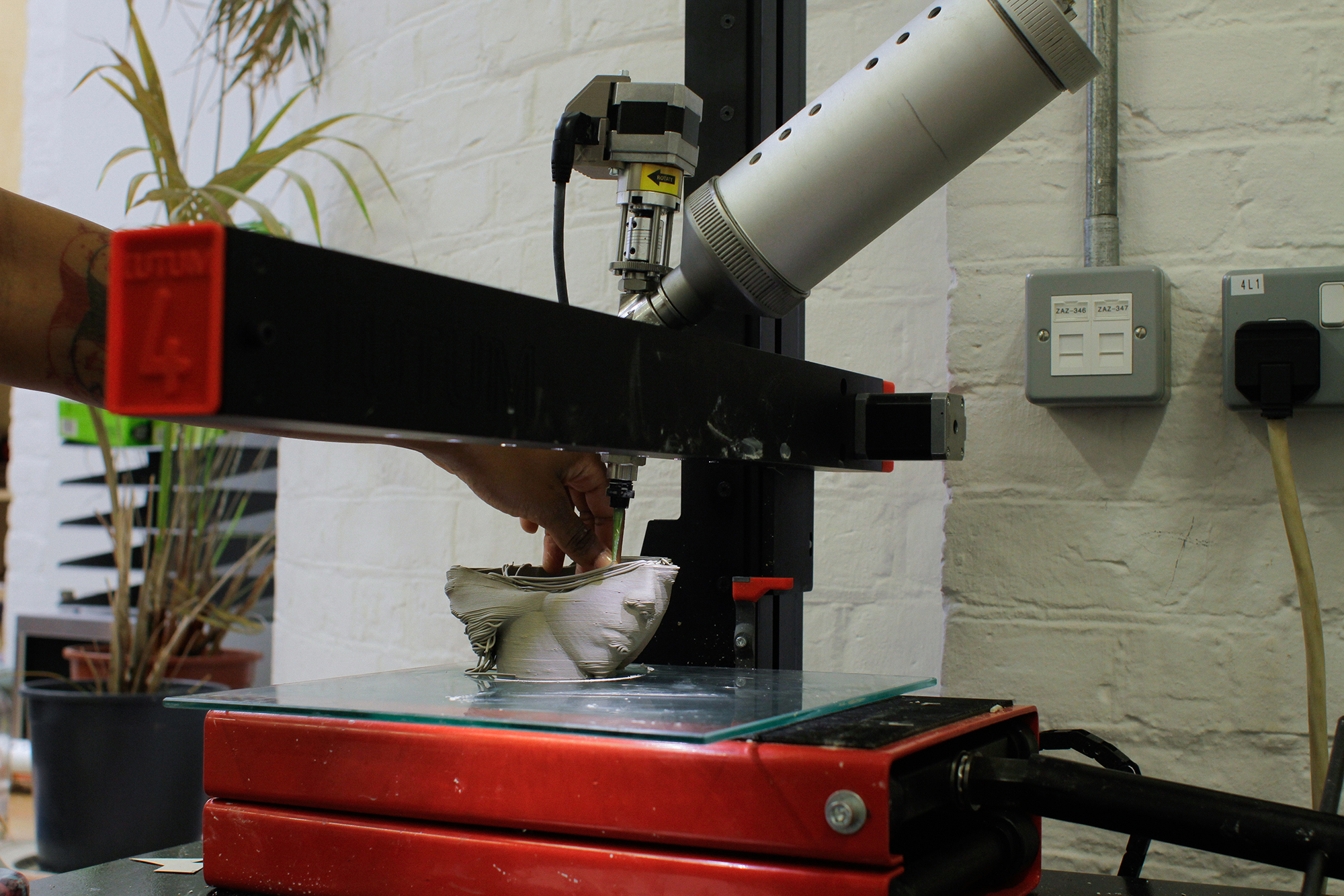 photo of a 3D ceramic printer on a desk, with a piece being printed midway and the artist's hand touching the piece from the left side. The piece on the printer shows two profile faces with closed eyes. Against a white walls with plants in the background on the left side